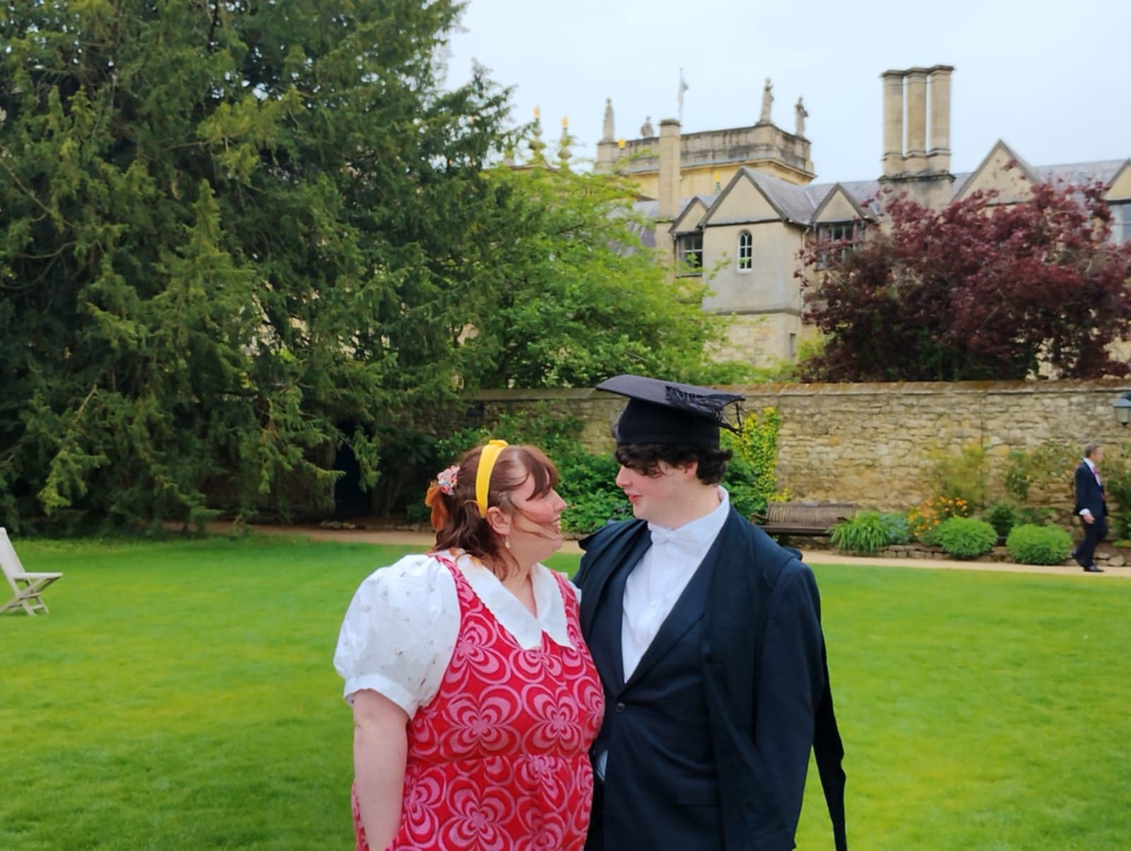 Finn and Rhianna looking at each other in the gardens of Trinity College, Oxford; Finn is dressed in academic dress